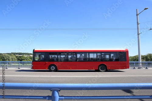 City bus crossing bridge. Red bus moving on road against blue barrier or guard rail.