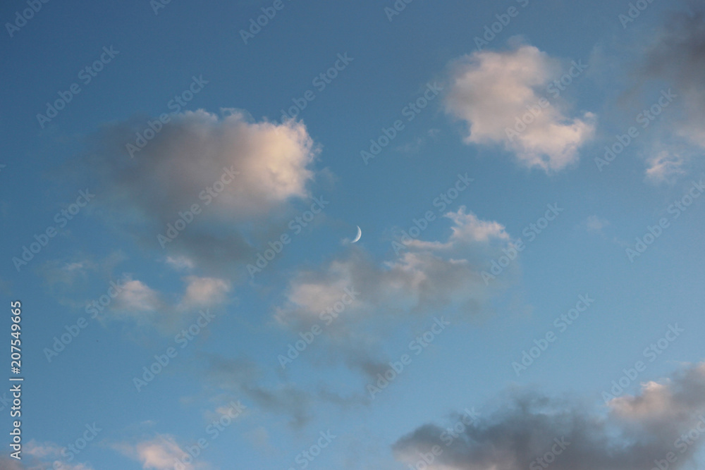 a thin crescent in the early evening is visible in the sky among the white clouds a thin crescent in the early evening is visible in the sky among the white clouds