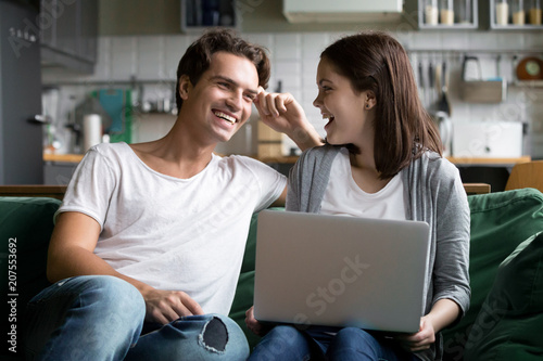 Happy millennial couple laughing joking with laptop together on kitchen sofa, smiling young girl and guy having fun talking at home on couch using computer, cheerful family enjoy weekend with device