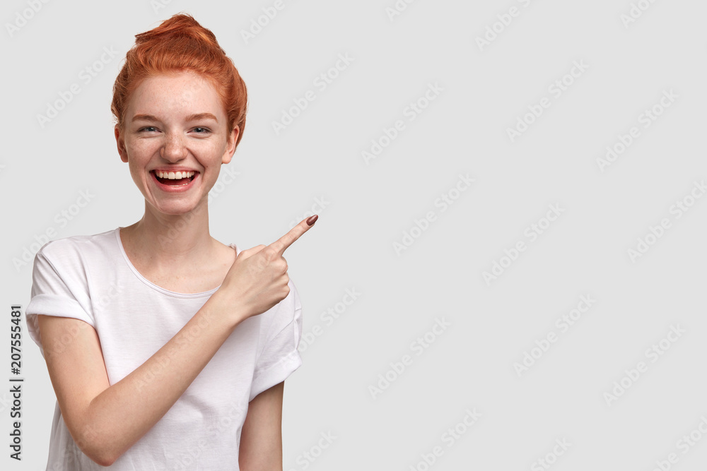 Positive tender freckled ginger young female with shining bright smile, dressed casually, indicates with fore finger at upper right corner, shows space for your advertisement or promotional text Positive tender freckled ginger young female with shining bright smile, dressed casually, indicates with fore finger at upper right corner, shows space for your advertisement or promotional text