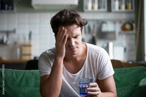 Young man suffering from strong headache or migraine sitting with glass of water in the kitchen, millennial guy feeling intoxication and pain touching aching head, morning after hangover concept photo