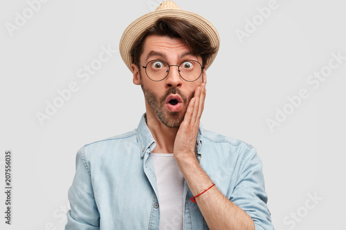Emotive shocked successful male farmer dressed in casual shirt and straw hat, stares at camera in bewilderment, keeps hand on cheek, poses against white studio background. Surprisement concept