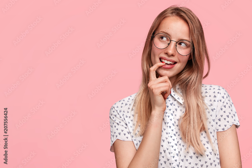 Glad positive young female looks thoughtfully and happily aside, keeps fore finger in mouth, remembers pleasant moment in life, stands against pink background with blank space for your hearder Glad positive young female looks thoughtfully and happily aside, keeps fore finger in mouth, remembers pleasant moment in life, stands against pink background with blank space for your hearder