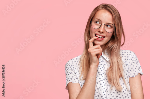 Glad positive young female looks thoughtfully and happily aside, keeps fore finger in mouth, remembers pleasant moment in life, stands against pink background with blank space for your hearder