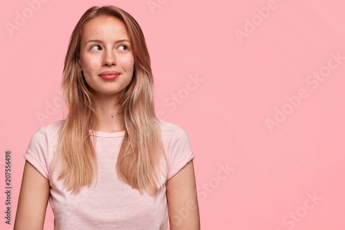 Beautiful cute young female with pensive look, concentrated upwards, has dreamy positive expression, thinks about something pleasant and unforgettable, poses against pink background. Copy space