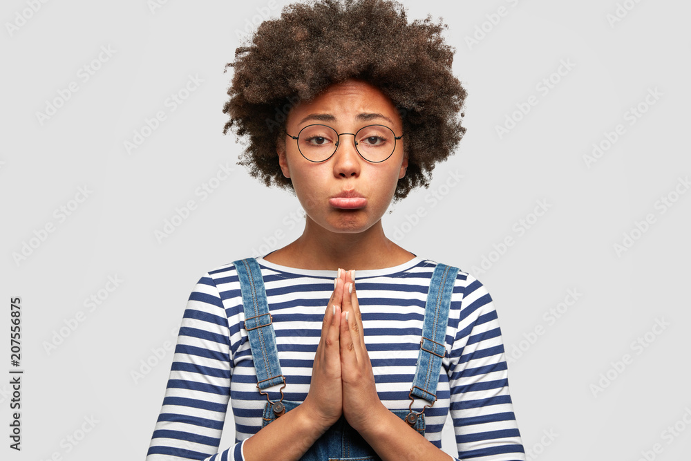 Unhappy gloomy African American female asks for something desirable, curves lower lip, keeps palms pressed together, has pleading expression. Lovely housewife feels guilty, tells: Forgive me Unhappy gloomy African American female asks for something desirable, curves lower lip, keeps palms pressed together, has pleading expression. Lovely housewife feels guilty, tells: Forgive me