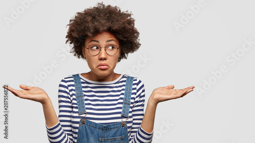 Cuadro en lienzo Hesitant puzzled young female with Afro haircut, shruggs shoulders and faces dilemma, looks clueless aside, keeps hands raised in palms, isolated on white background, makes difficult choice