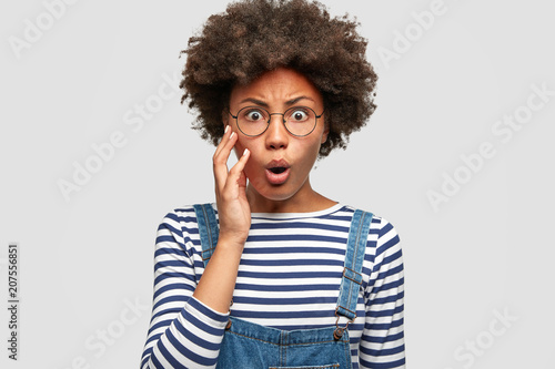 Photo of anxious African American female looks surprisingly and raises eyebrows in bewilderment, has Afro hairstyle, wears sriped jacket and denim dungarees, isolated over white studio wall.