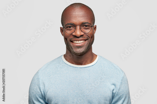 Middle aged cheerful dark skinned male with shining smile, wears light blue sweater, round spectacles, achieves everything by himself, stands against white studio wall, has relaxed face expression photo