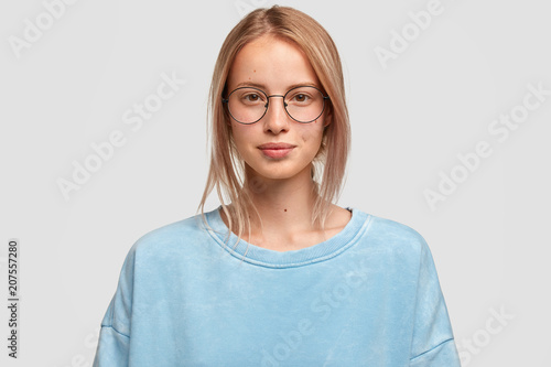 Headshot of attractive Caucasian female looks directly at camera, has no make up, healthy skin, wears big glasses and boyfriend`s sweater, spends spare time together with lover. Beauty, lifestyle