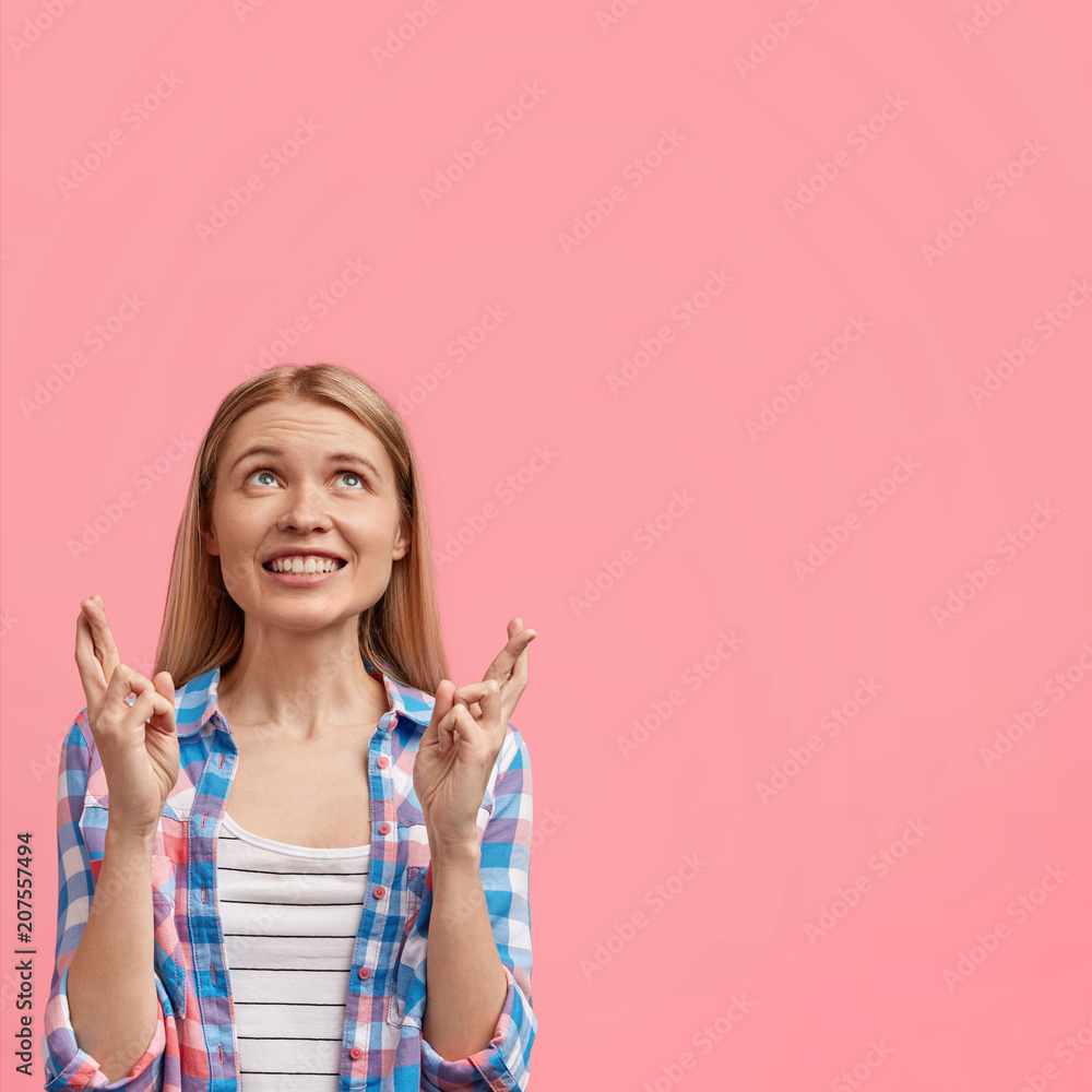 Young positive European female with broad warm smile, crosses fingers while makes wish, hopes dreams come true, wears casual checkered shirt, isolated over pink background with copy space aside Young positive European female with broad warm smile, crosses fingers while makes wish, hopes dreams come true, wears casual checkered shirt, isolated over pink background with copy space aside