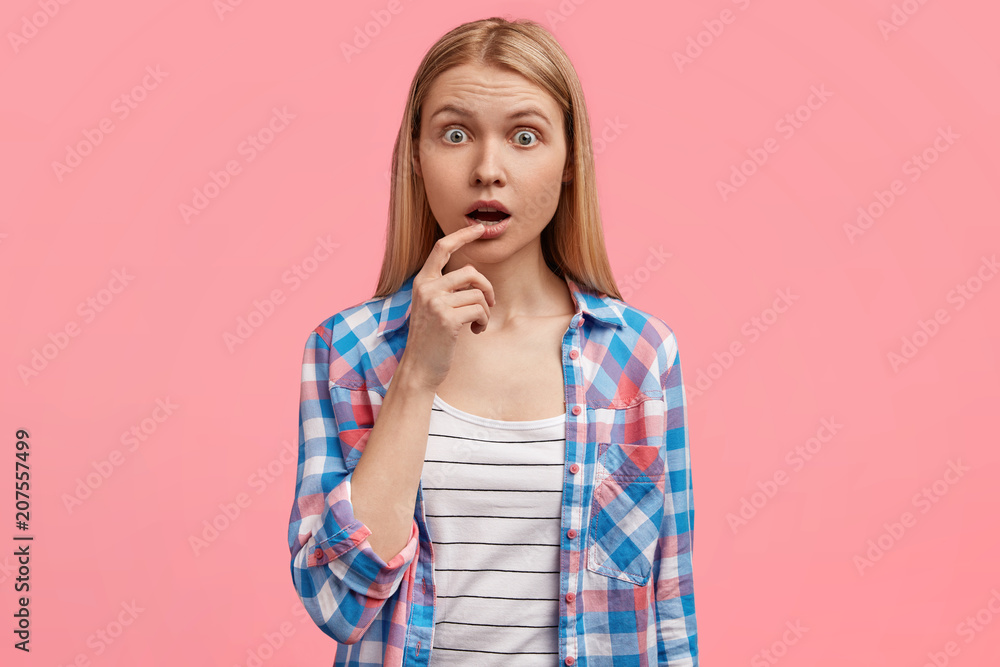 Portrait of astonished beautiful blonde European young woman looks surprisingly, keeps eyes popped out and opened mouth, wants to say something but being speechless, wears casual checkered shirt. Portrait of astonished beautiful blonde European young woman looks surprisingly, keeps eyes popped out and opened mouth, wants to say something but being speechless, wears casual checkered shirt.