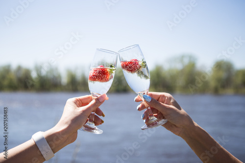 Two female hands holding a glass of champagne with strawberries. Background river