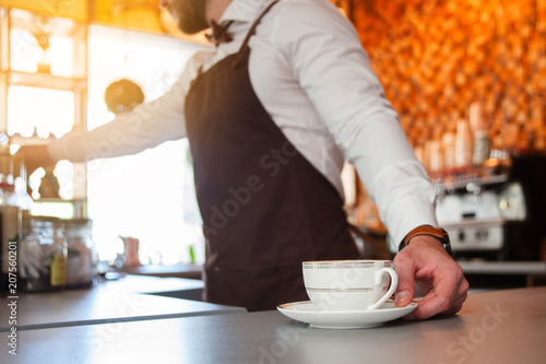 Cropped image of handsome barista in apron holding a cup of coffee at the bar counter in cafe. Cup in focus