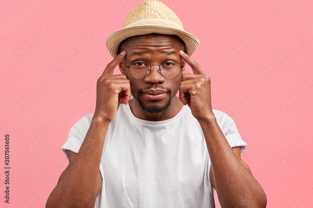 Curious dark skinned middle aged male keeps fore fingers on temples, wears straw hat, poses against pink background, tries to remember something important, has thoughtful expression, clever look Curious dark skinned middle aged male keeps fore fingers on temples, wears straw hat, poses against pink background, tries to remember something important, has thoughtful expression, clever look