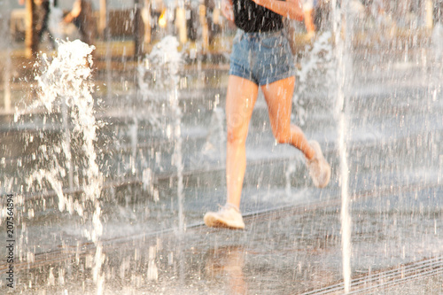 girl in shorts running in a city summer park between jets and spray of a fountain