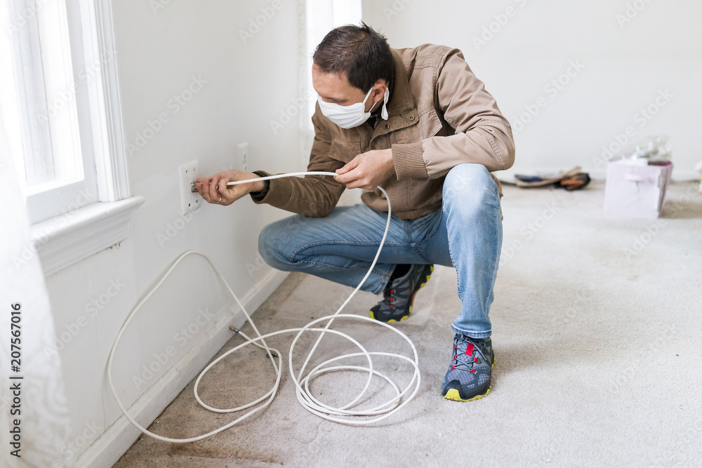 Young man in mask installing television cable internet wire in room wall carpet floor flooring, white walls, during remodeling renovation, cleaning, inspection Young man in mask installing television cable internet wire in room wall carpet floor flooring, white walls, during remodeling renovation, cleaning, inspection