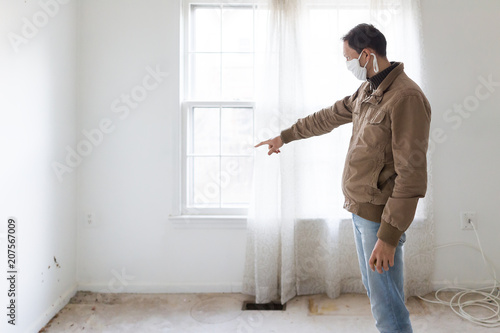 Young man in mask standing pointing at room wall carpet floor flooring, white painted walls, during remodeling renovation, cleaning, inspection of dirty mold, dust, trash in corner