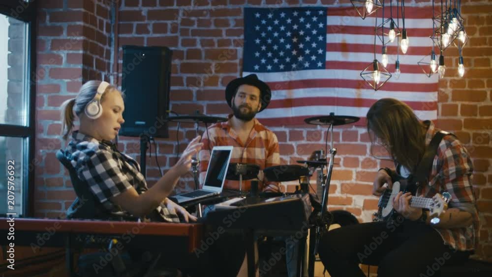 Cheerful blonde girl at synthesizer and men on drums and bass guitar creating music together in studio with American flag Cheerful blonde girl at synthesizer and men on drums and bass guitar creating music together in studio with American flag