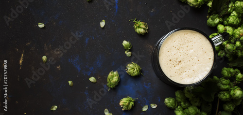 Big glass of foamy beer and hop cones on black stone background, top view