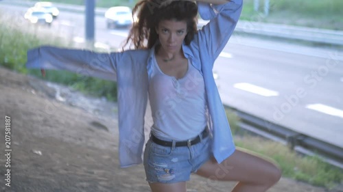 Curly dark hair girl in shorts and jeans shirt is dancing sensual under the road bridge 