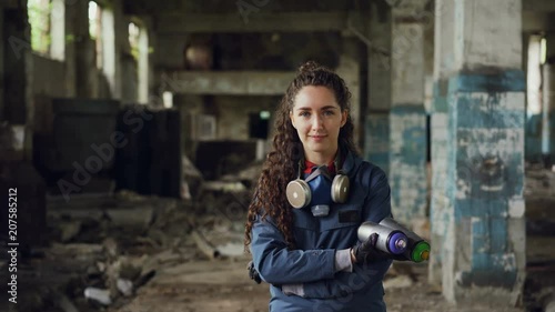 Portrait of smiling beautiful girl graffiti painter standing in old abandoned building, holding spray paints and looking at camera. Woman has protective mask and gloves.