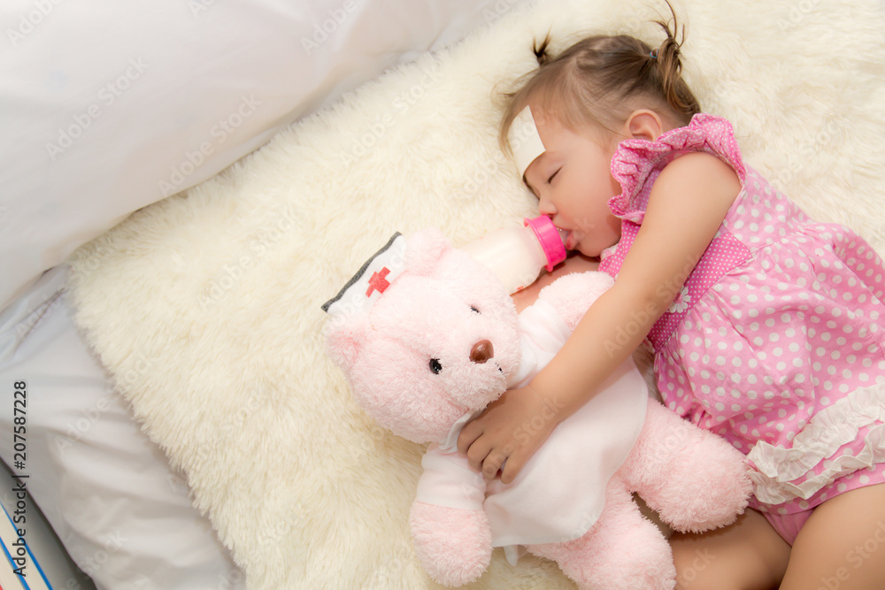 Baby drinking water or milk from a small bottle and lying on bed while hugging pink teddy bear in the bedroom because the baby having fever and attach cooling gel pad on her forehead for relief fever.
