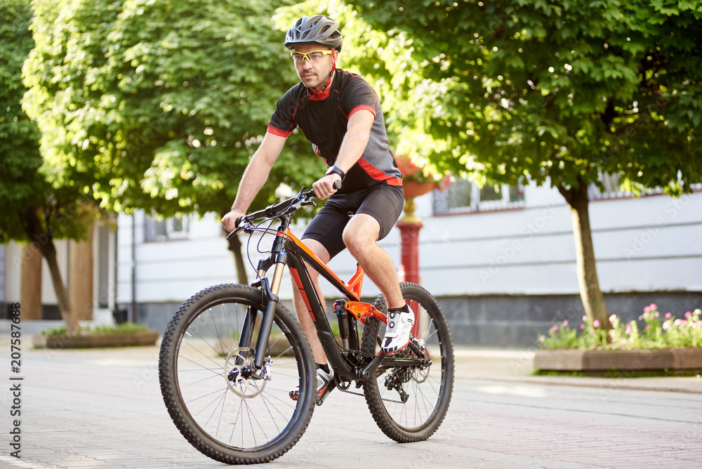 Obraz premium Athletic guy cyclist in cycling clothes and protective helmet, riding bike down empty city center alley surrounded by green trees. Sportsman training outdoors improving skills.