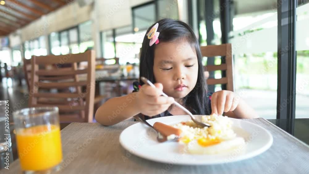 Asian children cute or kid girl enjoy eating fried rice with fried egg and sausage delicious food with orange juice on wood table for breakfast or lunch in cafe restaurant with holding spoon on slow Asian children cute or kid girl enjoy eating fried rice with fried egg and sausage delicious food with orange juice on wood table for breakfast or lunch in cafe restaurant with holding spoon on slow