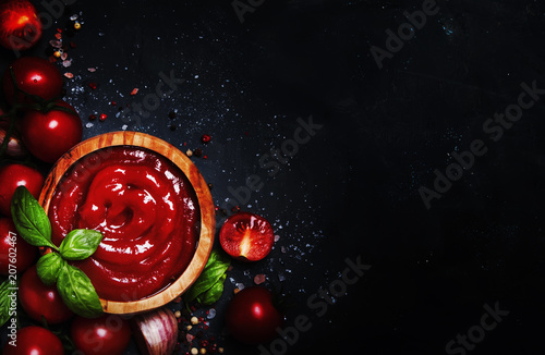 Hot tomato ketchup sauce with garlic, spices and green basil with cherry tomatoes in wooden bowl on gray stone background, top view