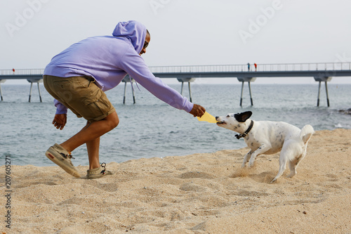 Active young African-American man hipster in sport hoody playing with his dog on the beach at sunny day