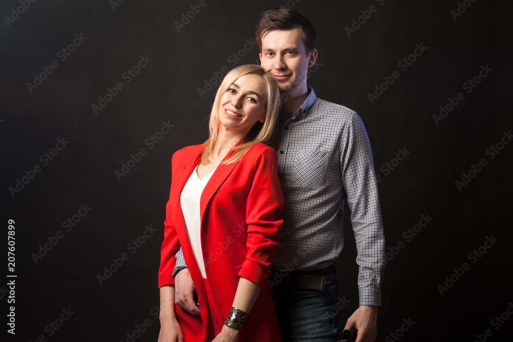 Obraz premium Young beautiful white blond girl in brightly red suit with jacket and white blouse and tall dark-haired guy hugging and kissing on isolated black background