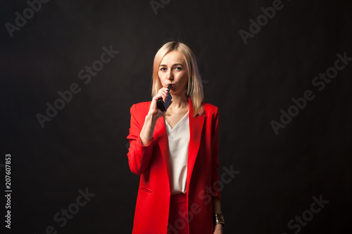Young beautiful white blond girl in a bright red strict suit with a jacket and white blouse looks at the camera and smoke vape on a black isolated background