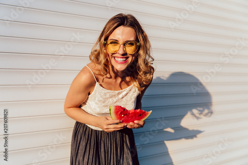 Excited girl, cheerfully smiling while looking at camera and eating a watermelon, wearing stylish clothes, yellow sunglasses, posing against the white wall, outdoors.