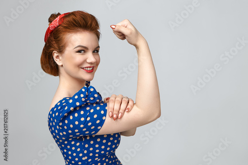 Isolated shot of charming positive young woman in retro clothes smiling broadly, tensing bicep, demonstrating her strong arms. Feminism, girl power, equal women's rights and independence concept photo