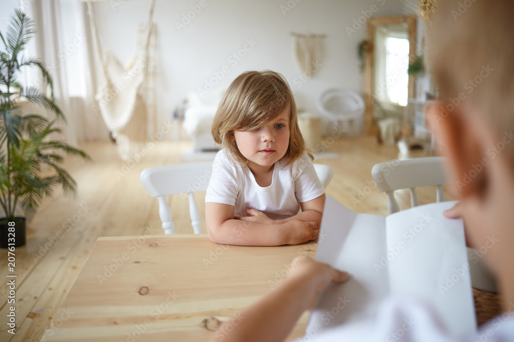 Indoor shot of charming cute little girl sitting at wooden table at home with her unrecognizable young father who is holding sheet of paper, checking her homework. Selective focus on kid's face Indoor shot of charming cute little girl sitting at wooden table at home with her unrecognizable young father who is holding sheet of paper, checking her homework. Selective focus on kid's face