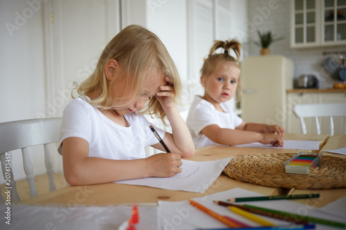 Cute blonde little boy doing homework, holding pen, drawing something on sheet of paper with his pretty baby sister sitting in background. Two children making drawings at wooden table in kitchen