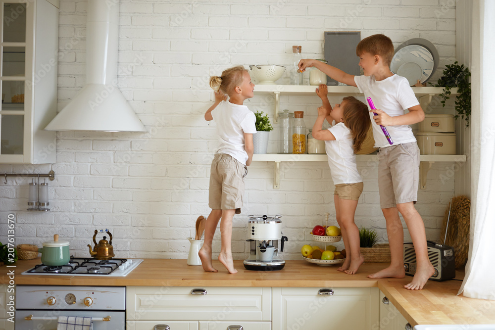 Happy childhood, family, fun and entertainment concept. Candid shot of three children siblings playing together in kitchen standing barefooted on wooden counter, looking for candies on shelf - obrazy, fototapety, plakaty 