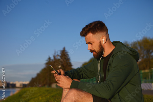 Outdoor shot of attractive young male man with stylish haircut and fuzzy beard enjoying nice summer weather, listening to music, sitting on grass by the river with earphones and mobile phone photo