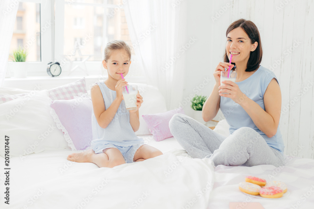 Cheerful mother and daughter dressed in pyjamas, have breakfast in morning, drink milk shake with doughnuts, sit crossed legs on comfortable bed in white bedroom. People and lifestyle concept