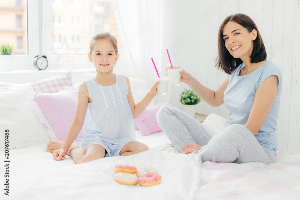 Portrait of happy young mother and her small daughter hold glasses of milk shake, have breakfast in bed, look positively at camera, enjoys weekend at home and togetherness, dressed in nightwear Portrait of happy young mother and her small daughter hold glasses of milk shake, have breakfast in bed, look positively at camera, enjoys weekend at home and togetherness, dressed in nightwear
