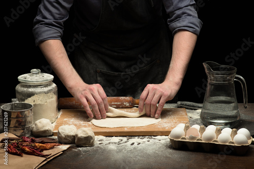 Chef hands are making a roll from dough on wooden board. black background