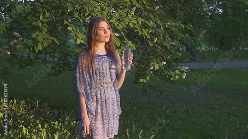 Beautiful female hiker drinking water in forest at sunset. A girl in a dress drinks cool water from a plastic bottle. photo
