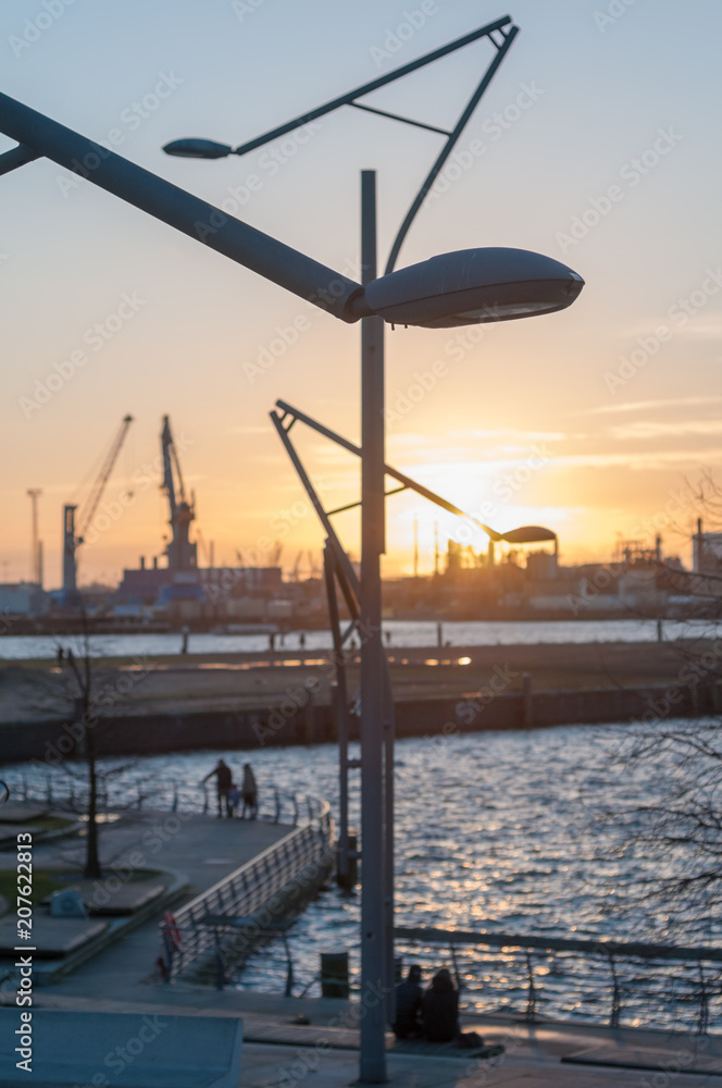 Abendszene mit Sonneuntergang bei den Marco-Polo-Terassen in Hafencity, Hamburg und Silhouetten von Menschen im unscharfen Hintergrund.