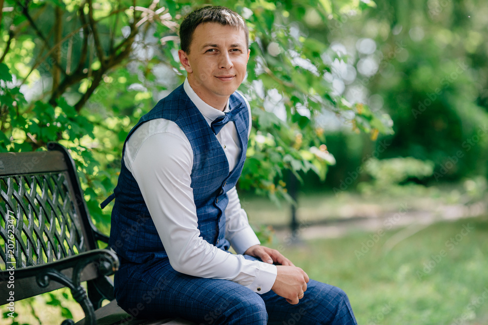 Portrait of a young man in a waistcoat and tight trousers in a cage. The groom sits on a bench in the street and enjoys thinking about the upcoming wedding, he wears a white shirt and a blue bow-tie. Portrait of a young man in a waistcoat and tight trousers in a cage. The groom sits on a bench in the street and enjoys thinking about the upcoming wedding, he wears a white shirt and a blue bow-tie.