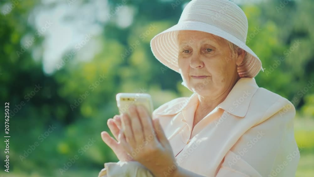 An elderly woman in summer clothes and a hat uses a smartphone. Relax in the summer park An elderly woman in summer clothes and a hat uses a smartphone. Relax in the summer park
