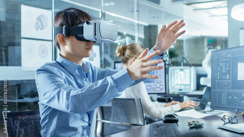 Computer Science Engineer wearing Virtual Reality Headset Works with 3D Modeling, Makes Gestures. In the Background Engineering Bureau with Busy Corowrkers. Computer Science Engineer wearing Virtual Reality Headset Works with 3D Modeling, Makes Gestures. In the Background Engineering Bureau with Busy Corowrkers.