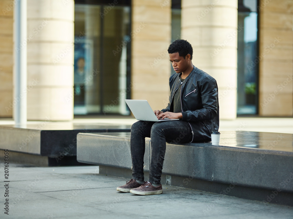 African American student in leather jacket sitting on stone bench outdoors and using laptop for studying African American student in leather jacket sitting on stone bench outdoors and using laptop for studying