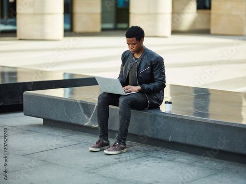 Young African man sitting on stone bench on city street and browsing Internet on laptop