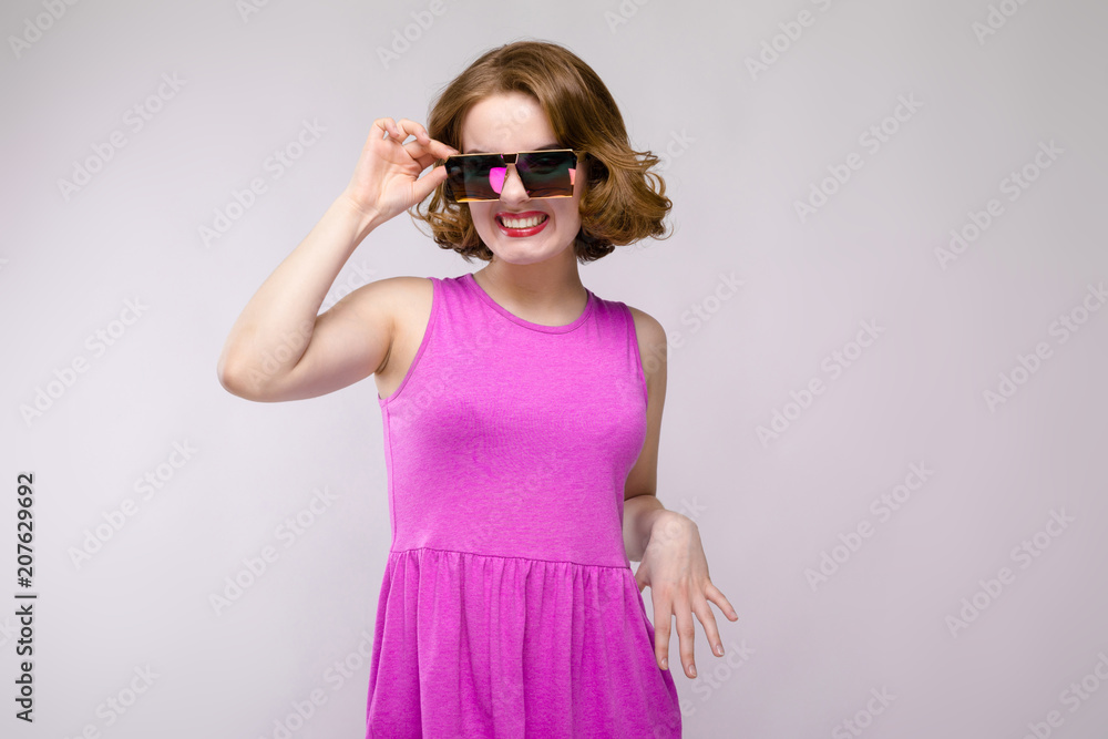 Charming young girl in pink dress on gray background. Cheerful girl in square glasses. The girl adjusts glasses Charming young girl in pink dress on gray background. Cheerful girl in square glasses. The girl adjusts glasses